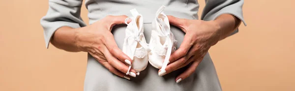 Panoramic shot of pregnant woman holding shoes isolated on beige — Stock Photo