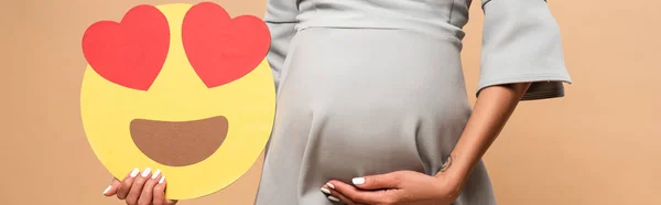 KYIV, UCRANIA - 1 de agosto de 2019: plano panorámico de una mujer embarazada sosteniendo sonriente con ojos de corazón sobre fondo beige - foto de stock