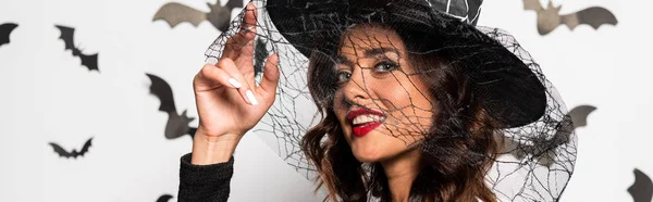 Панорамный снимок привлекательной женщины в шляпе ведьмы, смотрящей в камеру в Хэллоуин — стоковое фото