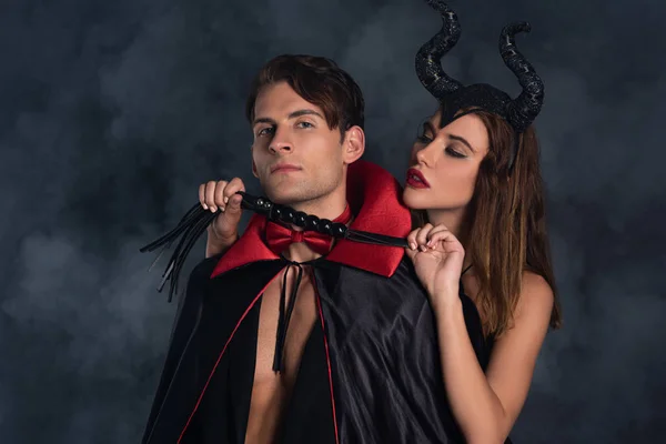 Attraente donna con le corna in possesso frusta fustigazione vicino all'uomo in costume di Halloween vampiro su nero con fumo — Foto stock