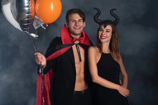 Cheerful man in vampire halloween costume holding balloons near girl with horns on black with smoke — Stock Photo