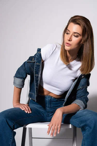 Fille élégante en jeans denim assis sur une chaise sur blanc — Photo de stock