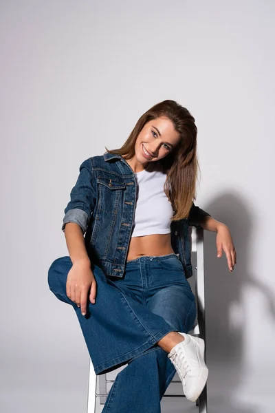 Cheerful girl in denim jeans sitting on chair on white — Stock Photo