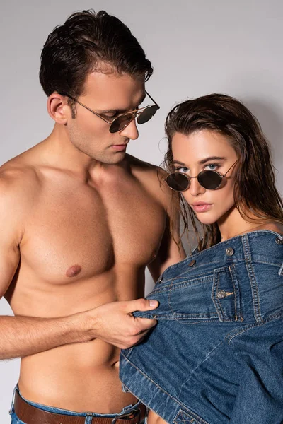 Stylish man in sunglasses touching denim jacket of girl on white — Stock Photo