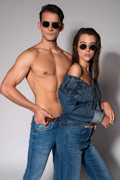 Uomo elegante e muscoloso in occhiali da sole toccare cintura vicino alla ragazza in giacca di jeans su bianco — Foto stock