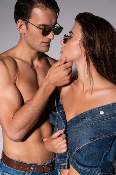 Стильный и мускулистый мужчина в солнцезащитных очках трогательное лицо девушки, изолированной на сером — стоковое фото