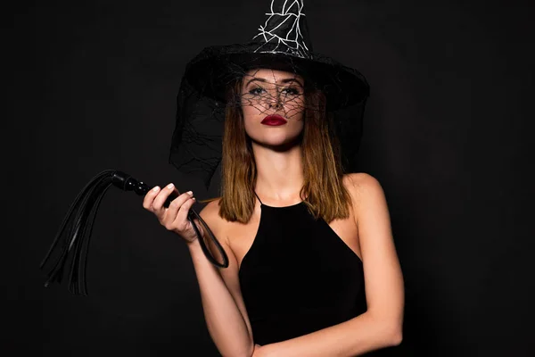 Mulher atraente em chapéu de bruxa segurando chicote açoitamento isolado no preto — Fotografia de Stock