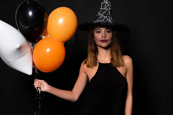 Young woman in witch hat holding balloons isolated on black — Stock Photo