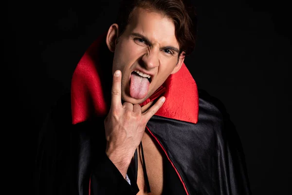Homme en vampire halloween costume collant sur la langue et montrant signe de roche isolé sur noir — Photo de stock