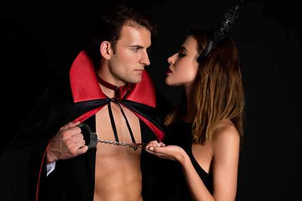 Hombre guapo en capa y mujer con cuernos sosteniendo esposas aisladas en negro - foto de stock