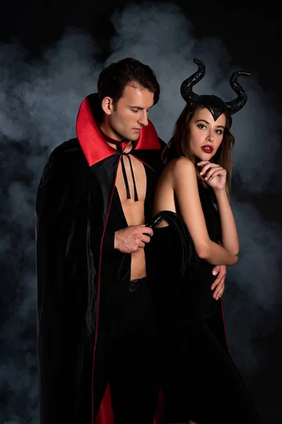 Handsome man in cloak holding flogging whip near woman with horns on black with smoke — Stock Photo