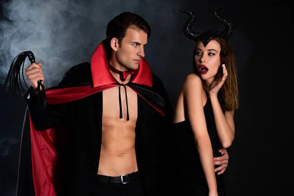 Handsome man in cloak holding flogging whip near surprised woman with horns on black with smoke — Stock Photo