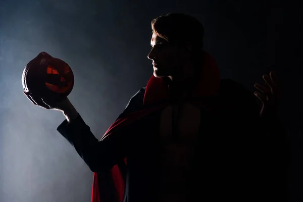 Handsome man holding spooky pumpkin and gesturing on black with smoke — Stock Photo