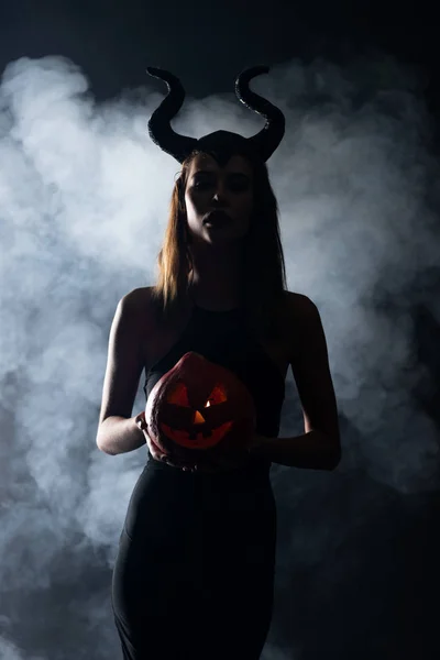 Silhouette of woman with horns holding spooky pumpkin on black with smoke — Stock Photo
