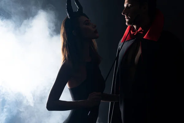 Handsome man in cloak looking at girl with horns holding flogging whip on black with smoke — Stock Photo