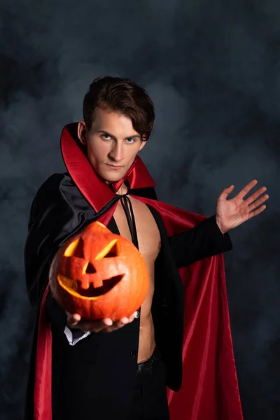 Handsome man holding halloween pumpkin and gesturing on black with smoke — Stock Photo