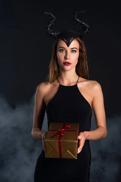 Attractive woman with horns holding present on black with smoke — Stock Photo