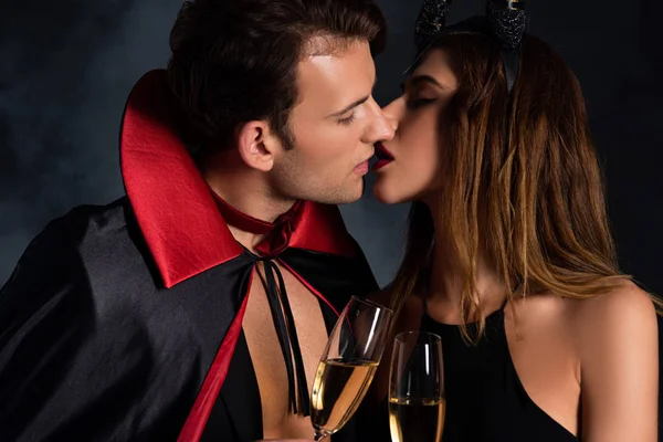 Homem bonito em capa e mulher com chifres beijando e segurando copos de champanhe em preto com fumaça — Fotografia de Stock