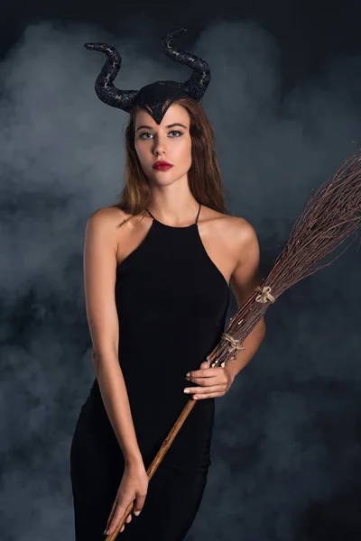 Jolie femme avec des cornes tenant balai d'Halloween sur noir avec de la fumée — Photo de stock