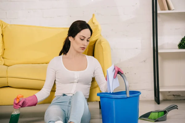 Femme au foyer fatiguée assis sur le sol et tenant bouteille de pulvérisation, seau et chiffon près de scoop et brosse — Photo de stock