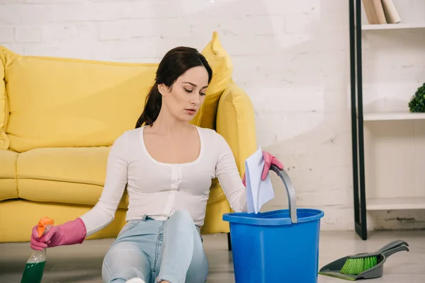 Tired housewife sitting on floor and holding spray bottle, bucket and rag near scoop and brush — Stock Photo