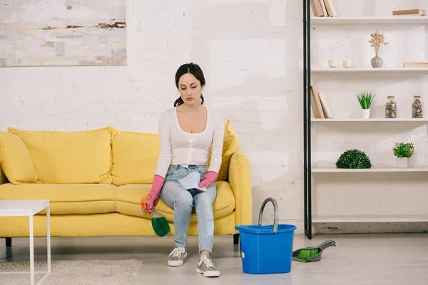 Müde Hausfrau sitzt auf gelbem Sofa und hält Sprühflasche neben Eimer, Schaufel und Bürste auf dem Boden — Stockfoto
