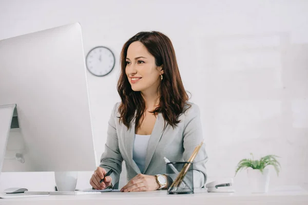 Attraktive, lächelnde Sekretärin, die auf Computermonitor schaut und in Notizbuch schreibt — Stockfoto