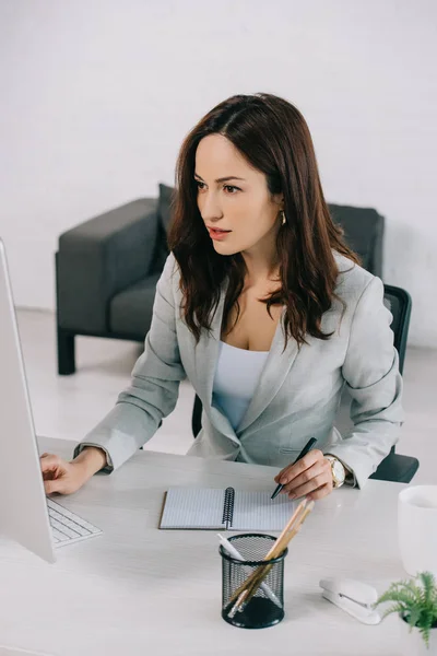 Secretaria atractiva y enfocada sentada en el lugar de trabajo y mirando el monitor de computadora — Stock Photo