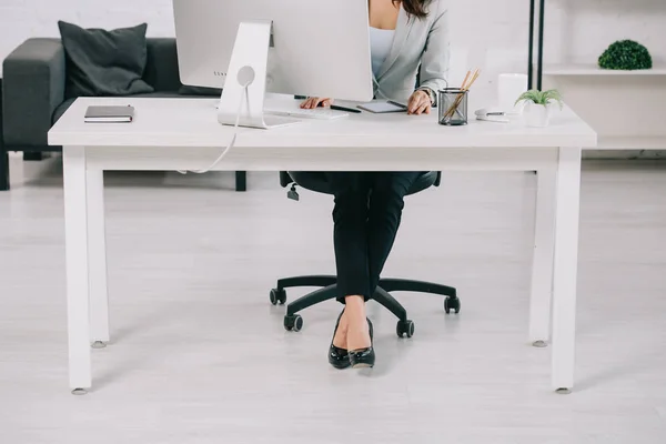 Vista patial del secretario en zapatos de tacón alto sentado en el lugar de trabajo en la silla de oficina - foto de stock