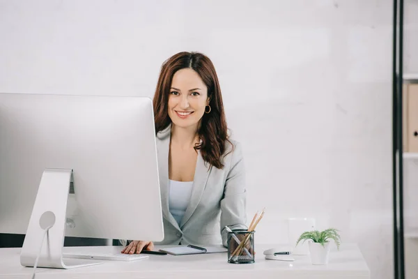 Attraktive, lächelnde Sekretärin, die am Arbeitsplatz in die Kamera schaut — Stockfoto