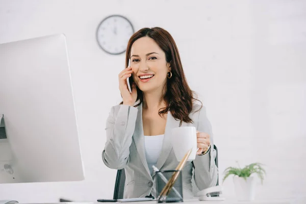 Alegre secretaria sosteniendo taza de café y hablando en el teléfono inteligente mientras está sentado en el lugar de trabajo - foto de stock