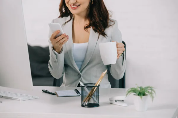 Vista recortada de la secretaria sonriente sosteniendo la taza de café y el uso de teléfono inteligente mientras está sentado en el lugar de trabajo - foto de stock