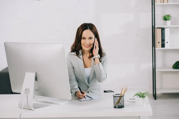 Smiling secretary talking on smartphone, looking at camera and writing in notebook while sitting at workplace — Stock Photo