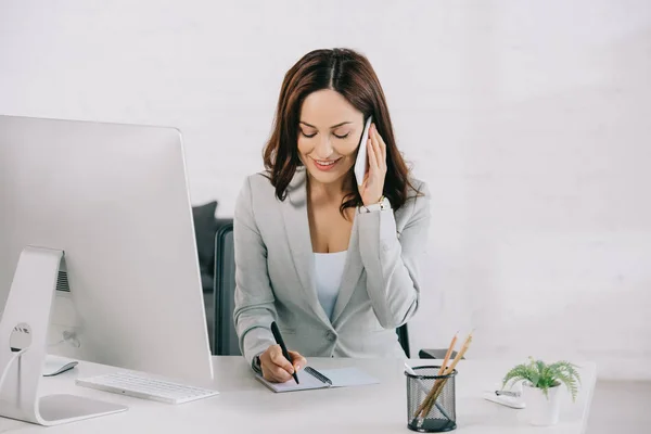 Secretaria sonriente hablando en el teléfono inteligente y escribiendo en el cuaderno mientras está sentado en el lugar de trabajo - foto de stock