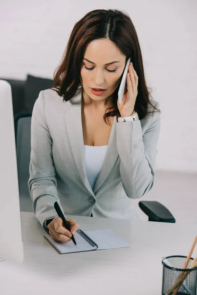 Attentive secretary talking on smartphone and writing in notebook while sitting at workplace — Stock Photo