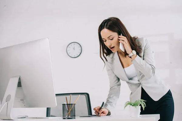 Attentive secretary talking on smartphone and writing in notebook while standing at workplace — Stock Photo