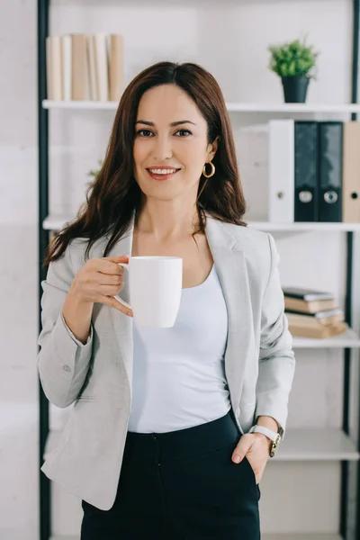 Hermosa, secretaria alegre mirando a la cámara mientras está de pie con la taza de café y sosteniendo la mano en el bolsillo - foto de stock