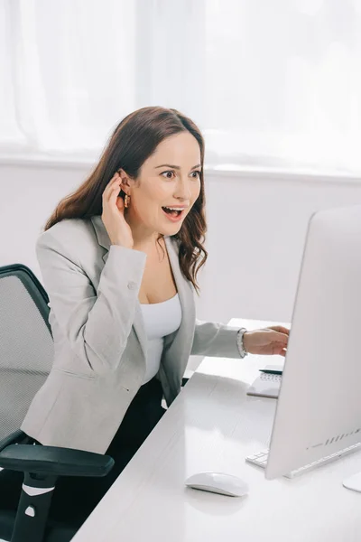 Secretaria emocionada y sorprendida mirando el monitor de la computadora mientras está sentada en el lugar de trabajo — Stock Photo