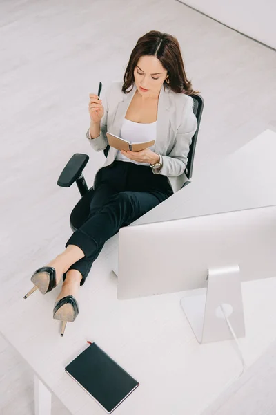 Vista de ángulo alto de la secretaria joven y concentrada mirando el cuaderno mientras está sentada en el lugar de trabajo - foto de stock