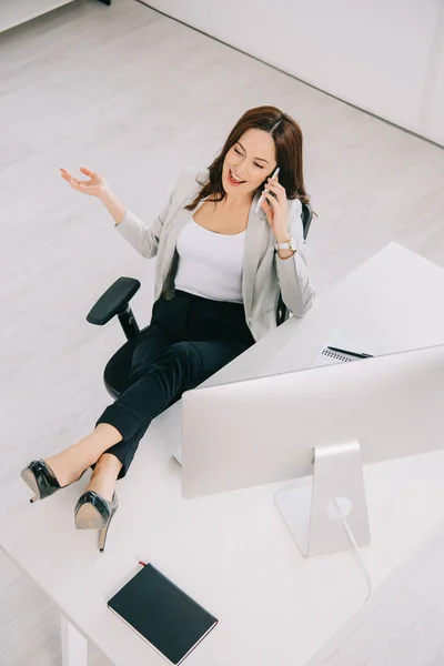 Vista de ángulo alto de alegre, elegante secretaria hablando en el teléfono inteligente mientras está sentado en el lugar de trabajo - foto de stock