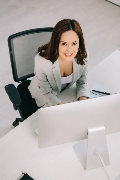 Vista de ángulo alto de la secretaria joven y sonriente mirando a la cámara mientras está sentada en el lugar de trabajo - foto de stock