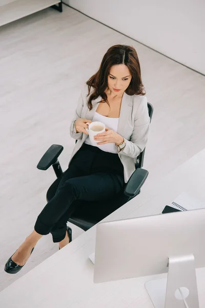 Blick aus der Höhe auf eine junge, elegante Sekretärin, die in der Nähe ihres Arbeitsplatzes sitzt und eine Kaffeetasse in der Hand hält — Stockfoto