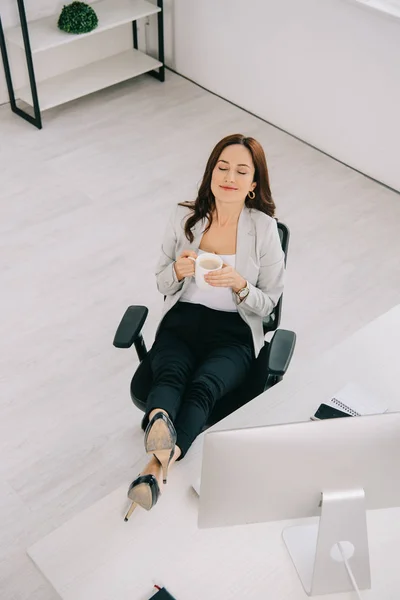 Vista de ángulo alto de la secretaria feliz y elegante sentada cerca del lugar de trabajo con los ojos cerrados y sosteniendo la taza de café - foto de stock