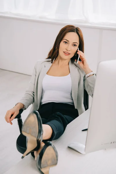 Nachdenkliche, elegante Sekretärin, die mit dem Smartphone spricht, während sie mit den Beinen auf dem Schreibtisch sitzt — Stockfoto