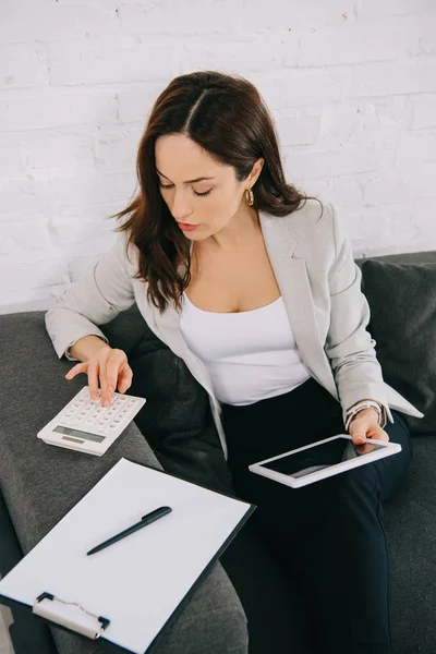 Concentrated young secretary using calculator and holding digital tablet while sitting on couch in office — Stock Photo