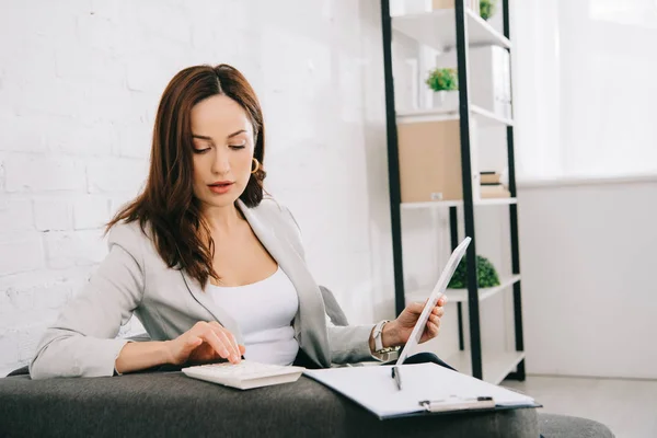 Attentive young secretary using calculator and holding digital tablet while sitting on couch in office — Stock Photo