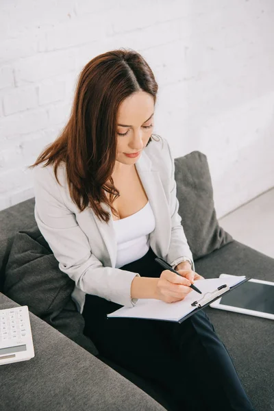 Attentive young secretary writing on clipboard while sitting on couch in office — Stock Photo