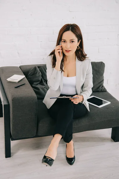 Aufmerksame junge Sekretärin spricht auf dem Smartphone, während sie auf der Couch neben dem digitalen Tablet sitzt — Stockfoto