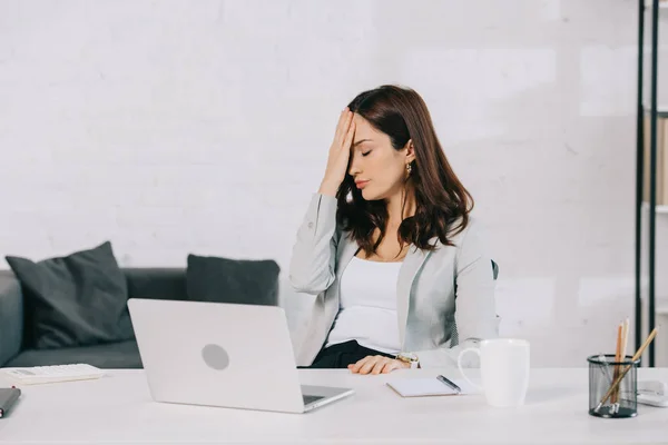 Erschöpfte Sekretärin leidet unter Kopfschmerzen, während sie am Arbeitsplatz sitzt und die Hand am Kopf hält — Stockfoto