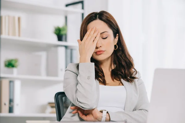 Exhausted secretary suffering from headache while sitting with closed eyes and holding hand near head — Stock Photo